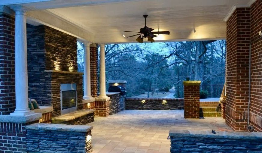 Covered stone patio with brick columns, ceiling fan, and a stone fireplace overlooking a snowy yard