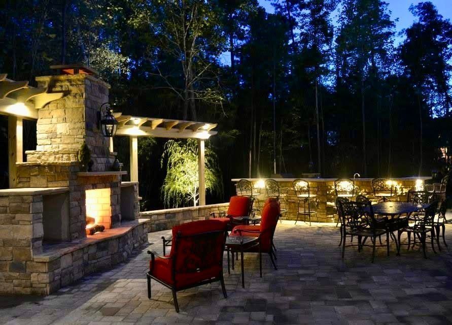 Stone patio with red chairs and tables beside an outdoor fireplace at dusk in a wooded setting