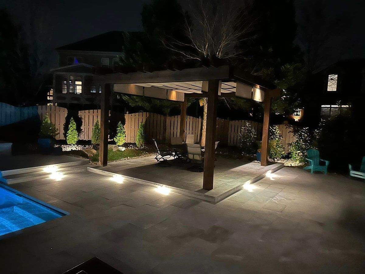 Nighttime backyard patio with a lit pergola, pool edge, and landscaped seating area