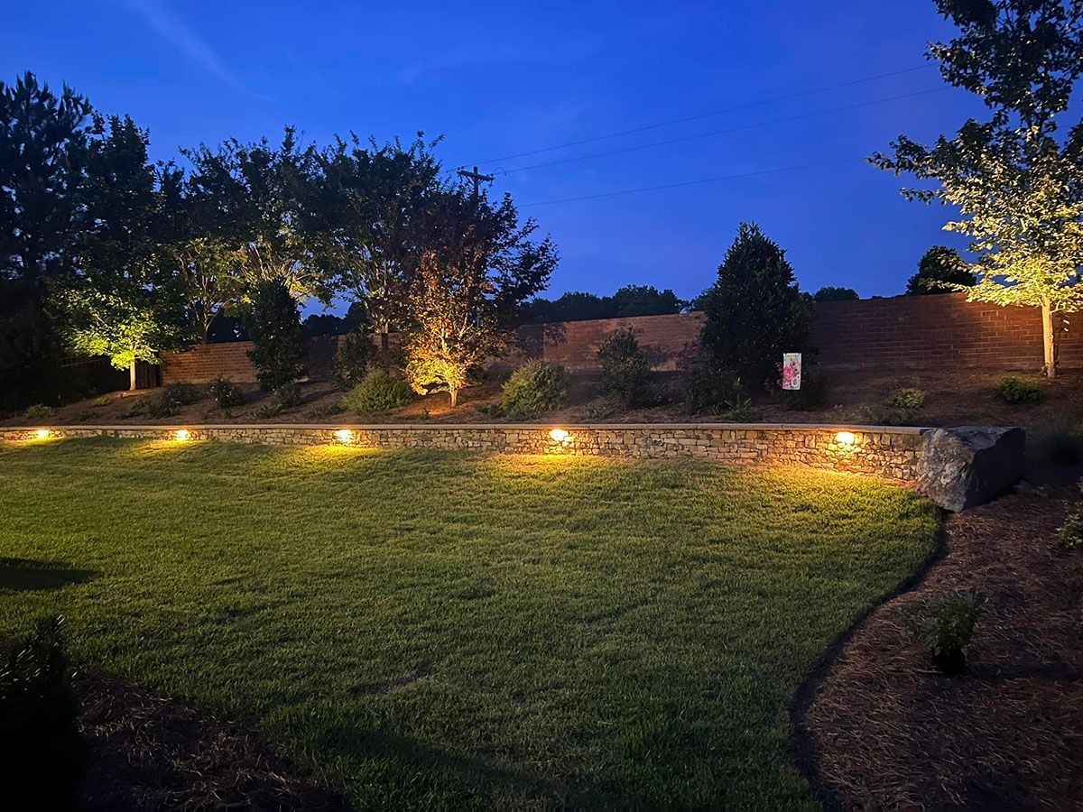 Lit garden path at dusk with trees, stone wall, and warm landscape lights under a blue sky