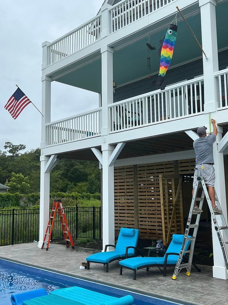 Poolside deck with white railings, American flag, rainbow windsock, and a person on a ladder painting.
