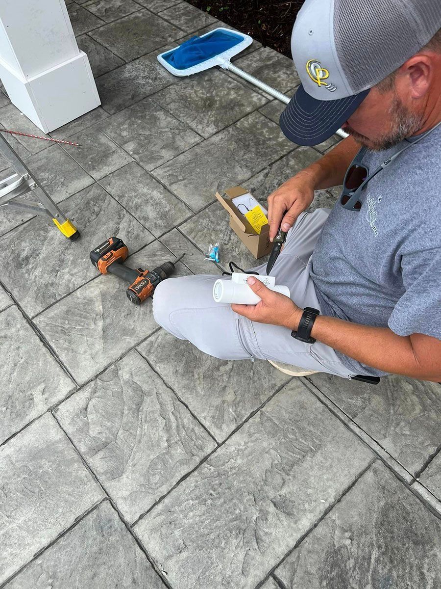 Man kneeling on a patio, working with tools and wires beside a blue dustpan and paver stones