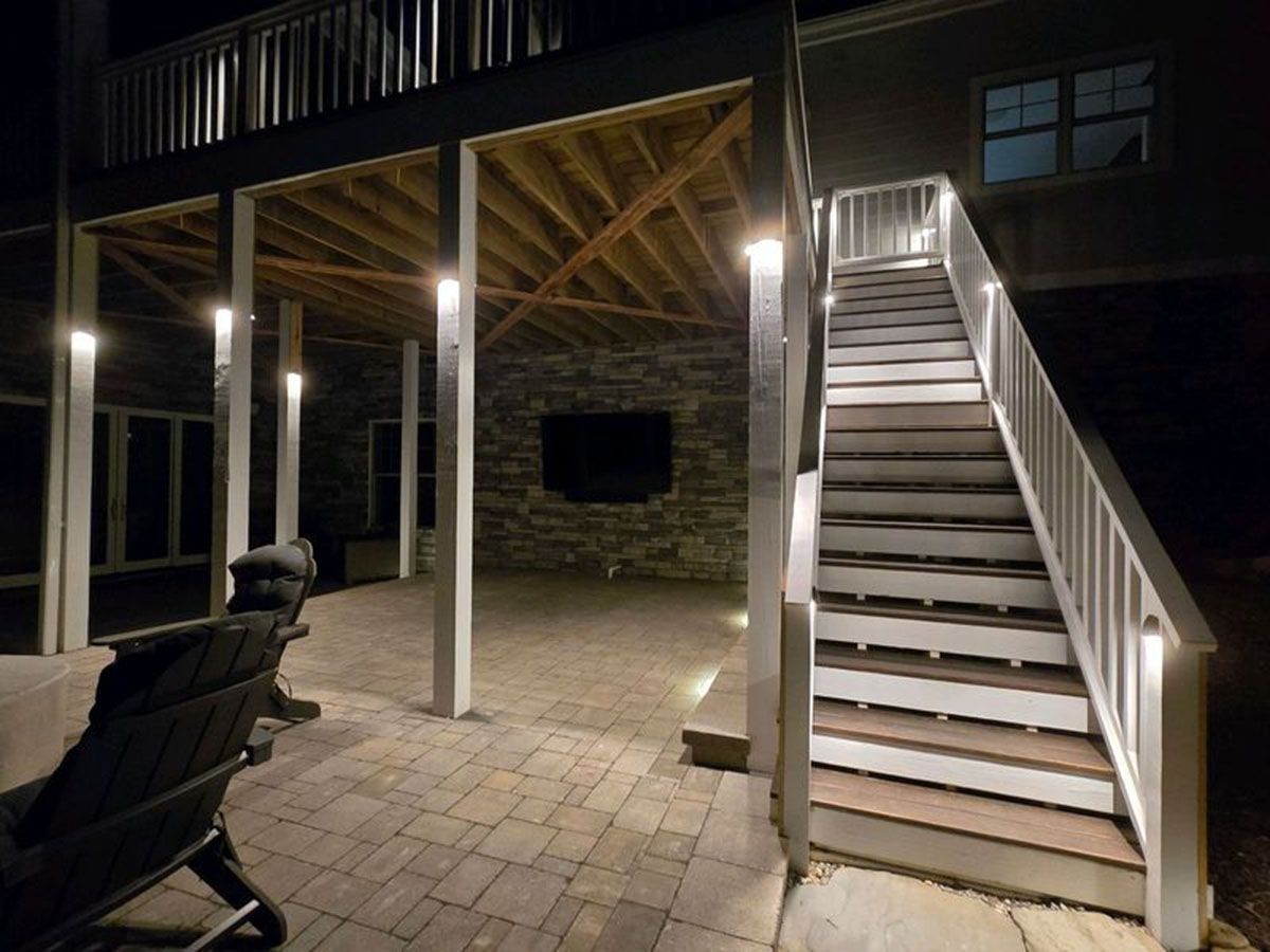 Covered backyard patio at night with stone pavers, string lights, and an exterior staircase to an upper deck
