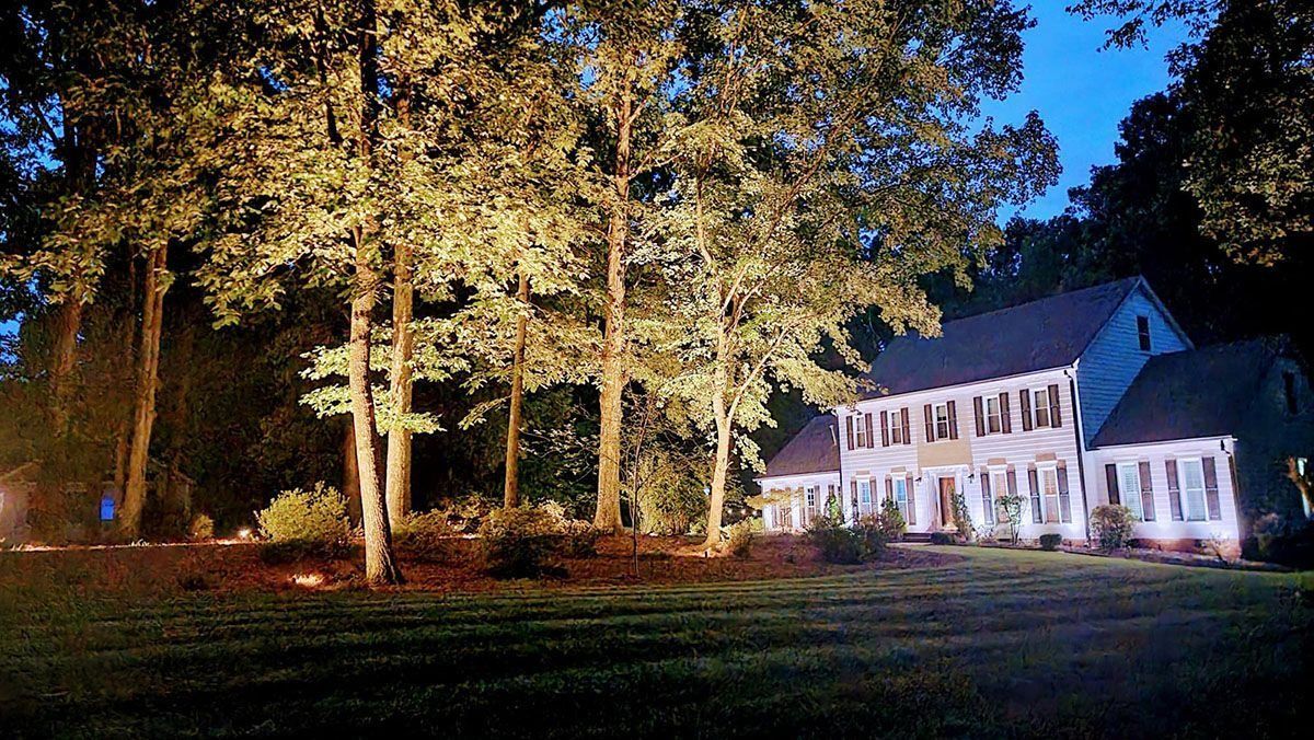 White two-story house at night beside tall trees, lit by bright exterior lights.