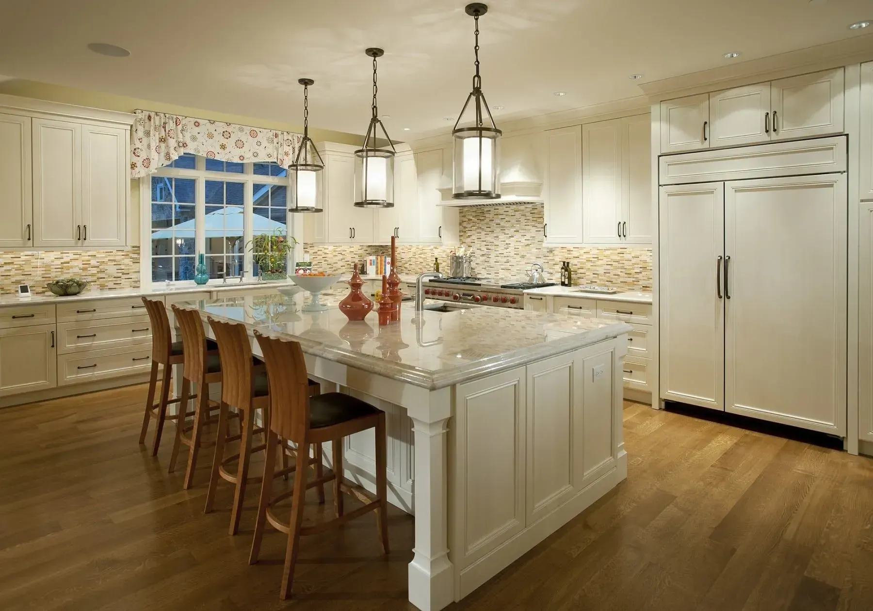 Elegant white kitchen with island, pendant lights, wooden floors, and tall refrigerator.