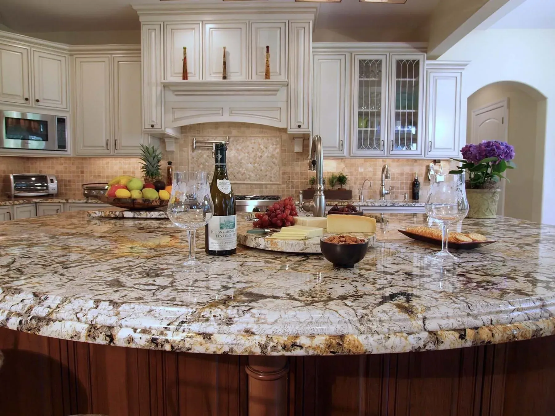 Elegant kitchen with granite countertop, wine, cheese, and fruit on the island, white cabinets.