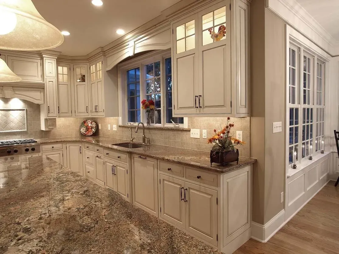 Cream-colored kitchen cabinets with granite countertops, sink, and large windows. Warm lighting and decor.