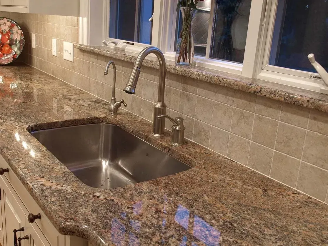 Stainless steel sink in a kitchen with granite countertops and a window.