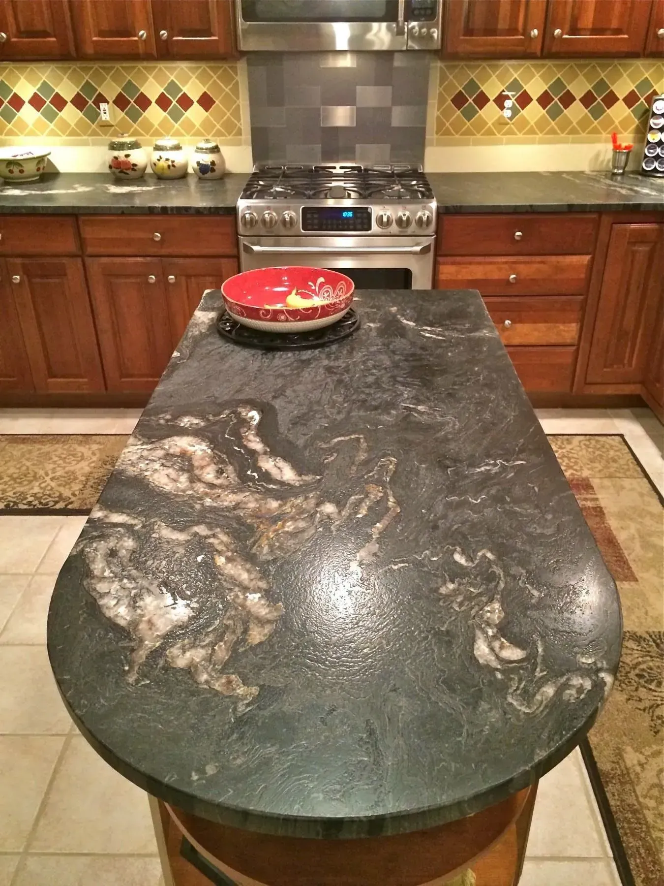 Kitchen island with a dark, marbled countertop and a red bowl, stainless steel range in background.