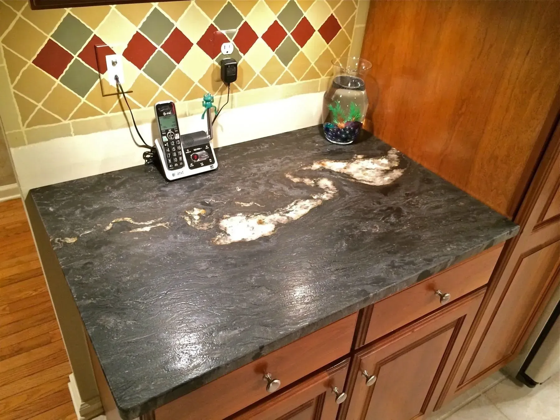 Kitchen countertop with dark granite, brown cabinets, and colorful backsplash tiles.