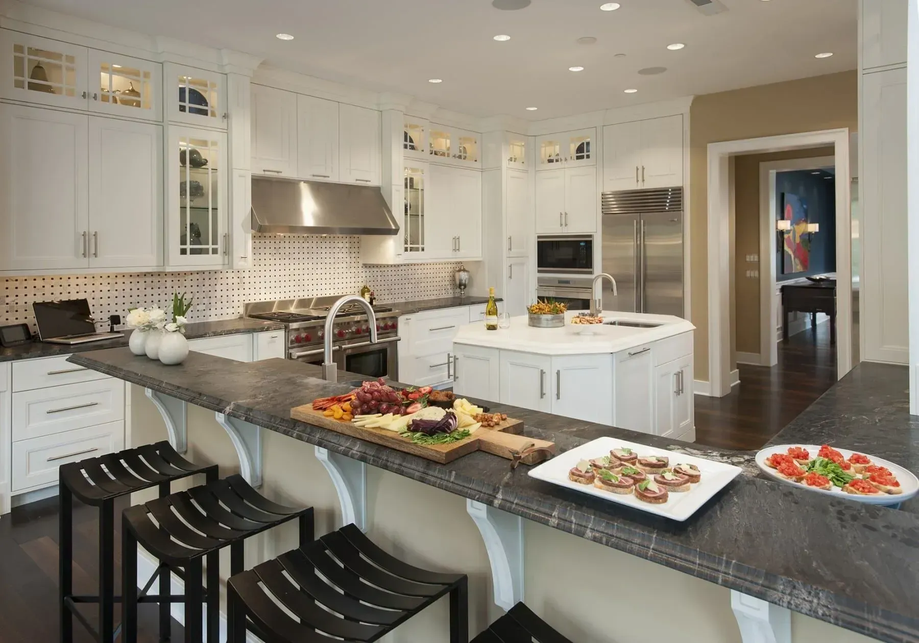 White kitchen with dark countertops, island, and bar seating. Food platters are arranged on the bar.