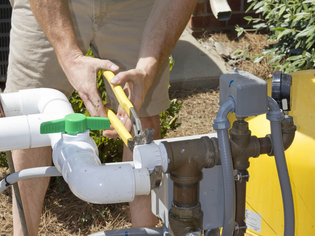 Hands using yellow pliers to work on a white PVC pipe valve outdoors beside a yellow tank.