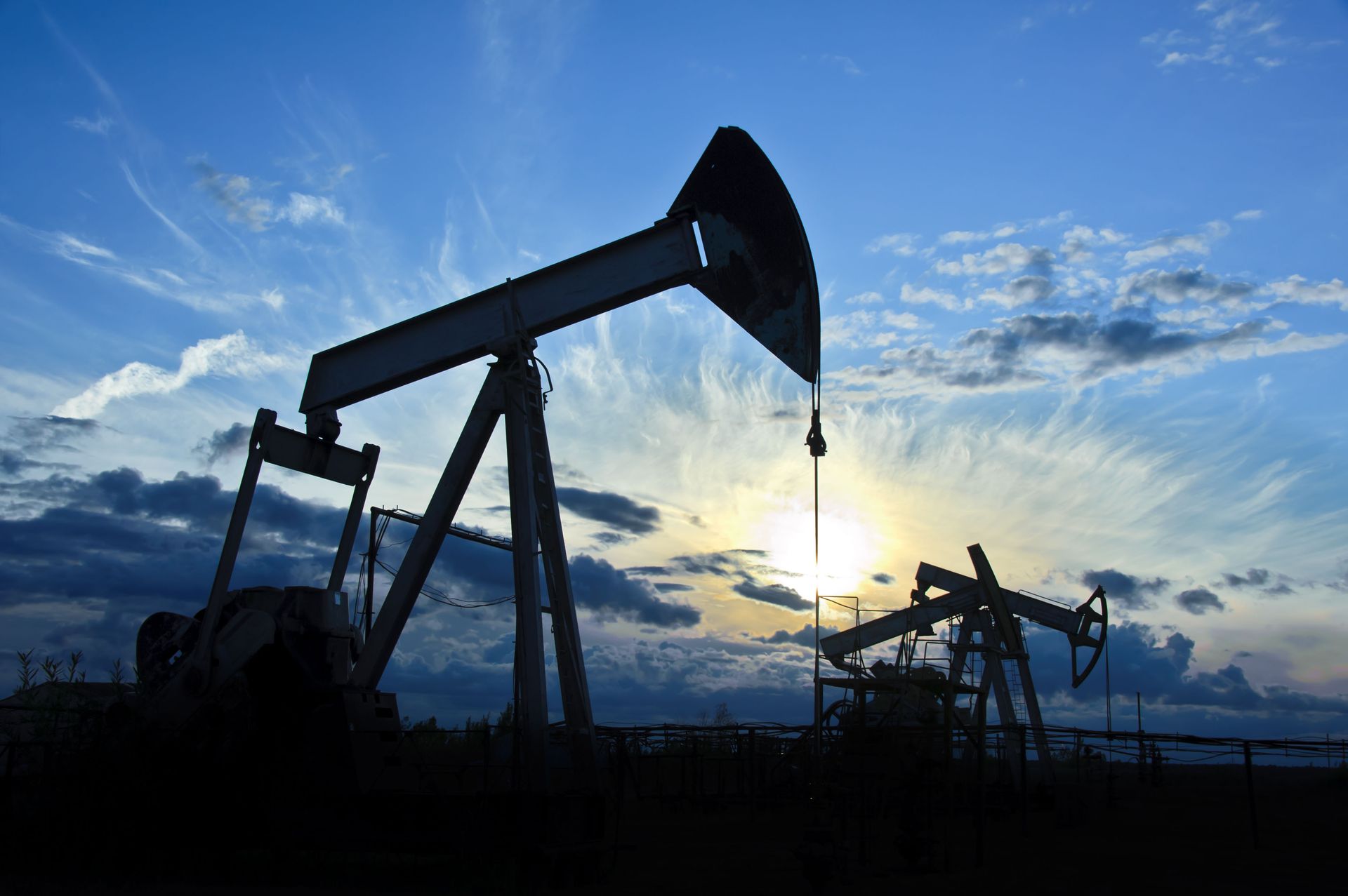Silhouetted oil pumpjacks at sunset beneath a blue sky with clouds