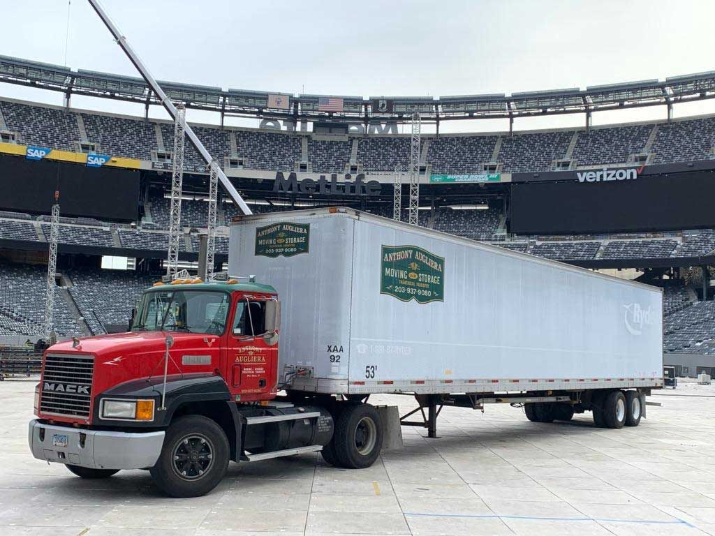 A red semi truck with a white trailer is parked in front of a stadium.