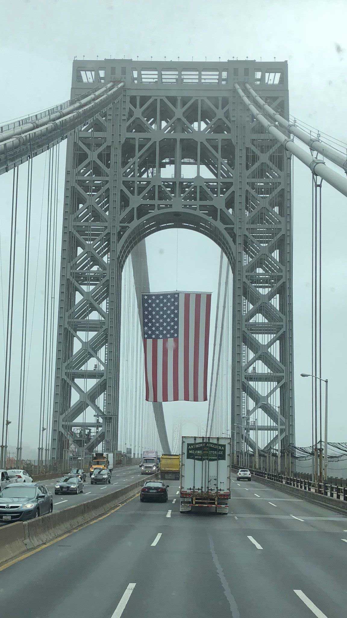 A truck is driving under a bridge with an american flag hanging from it.