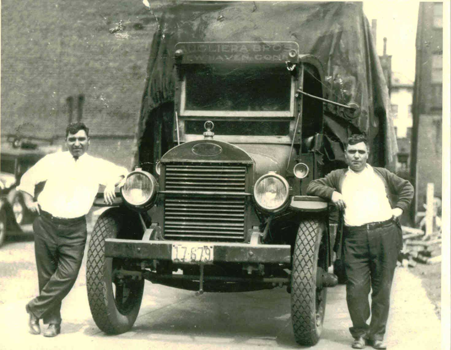 A black and white photo of two men standing in front of an old truck