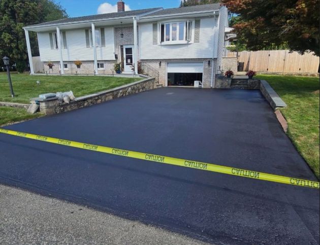 A newly paved asphalt driveway leads to a single-story white house with a garage, blocked off by yellow caution tape.
