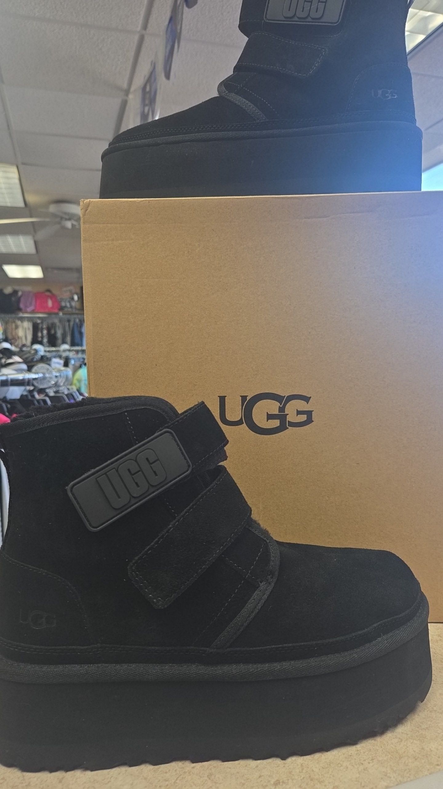 Black UGG platform boots with Velcro straps, sitting on top of an UGG box, retail setting.