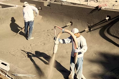 Two workers applying concrete to a pool construction site. One uses a hose, the other levels the surface.