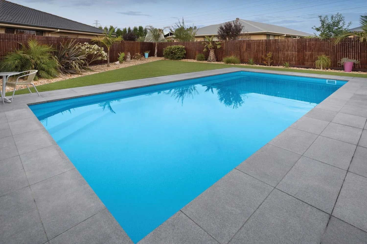 Rectangular swimming pool with blue water, surrounded by gray tiles, green grass, and a wooden fence.