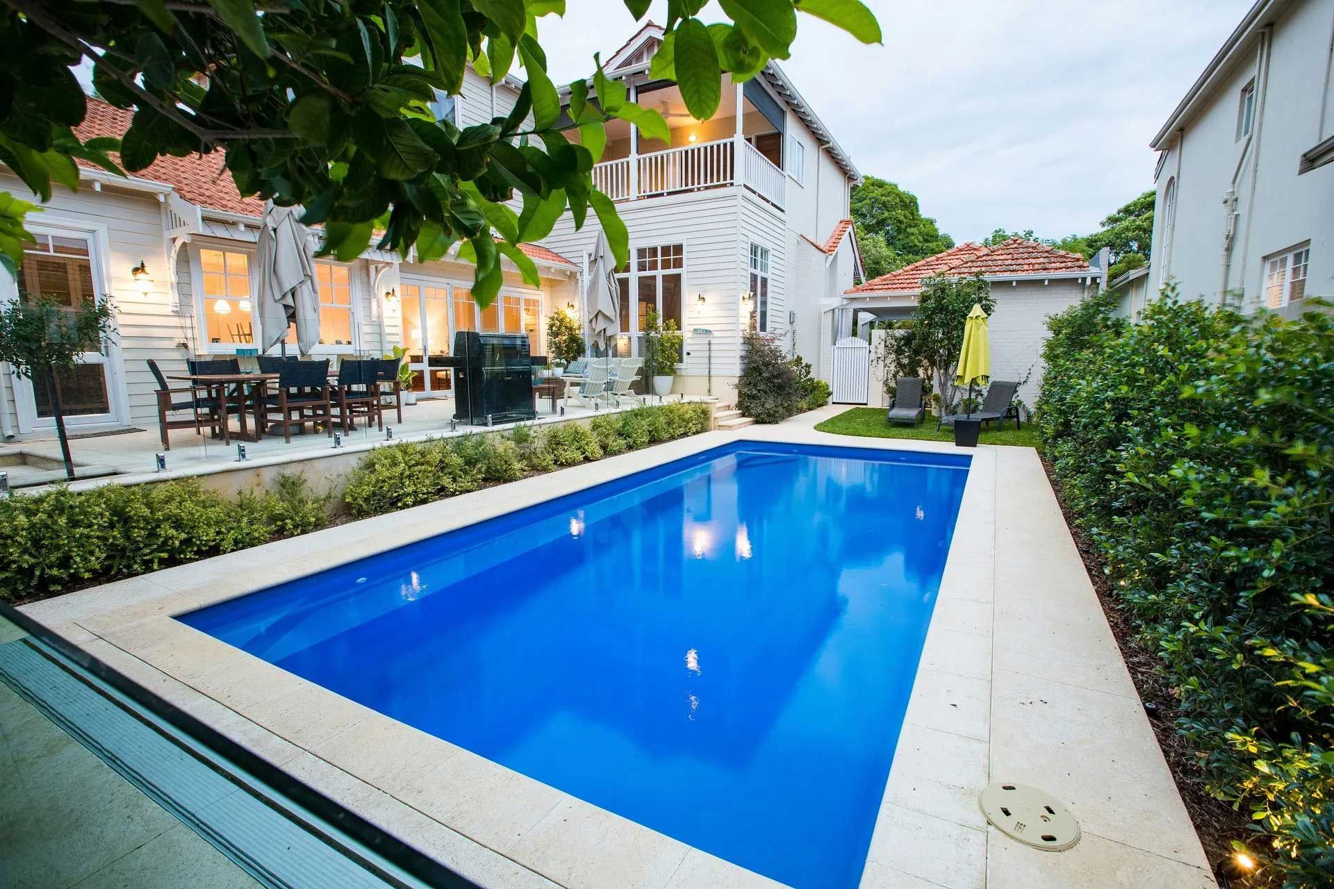 A backyard with a blue rectangular pool, a white house, and greenery.