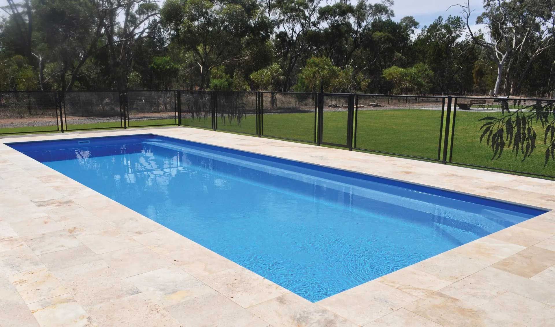 Rectangular blue swimming pool with tan tile surround, black fence, and green lawn.