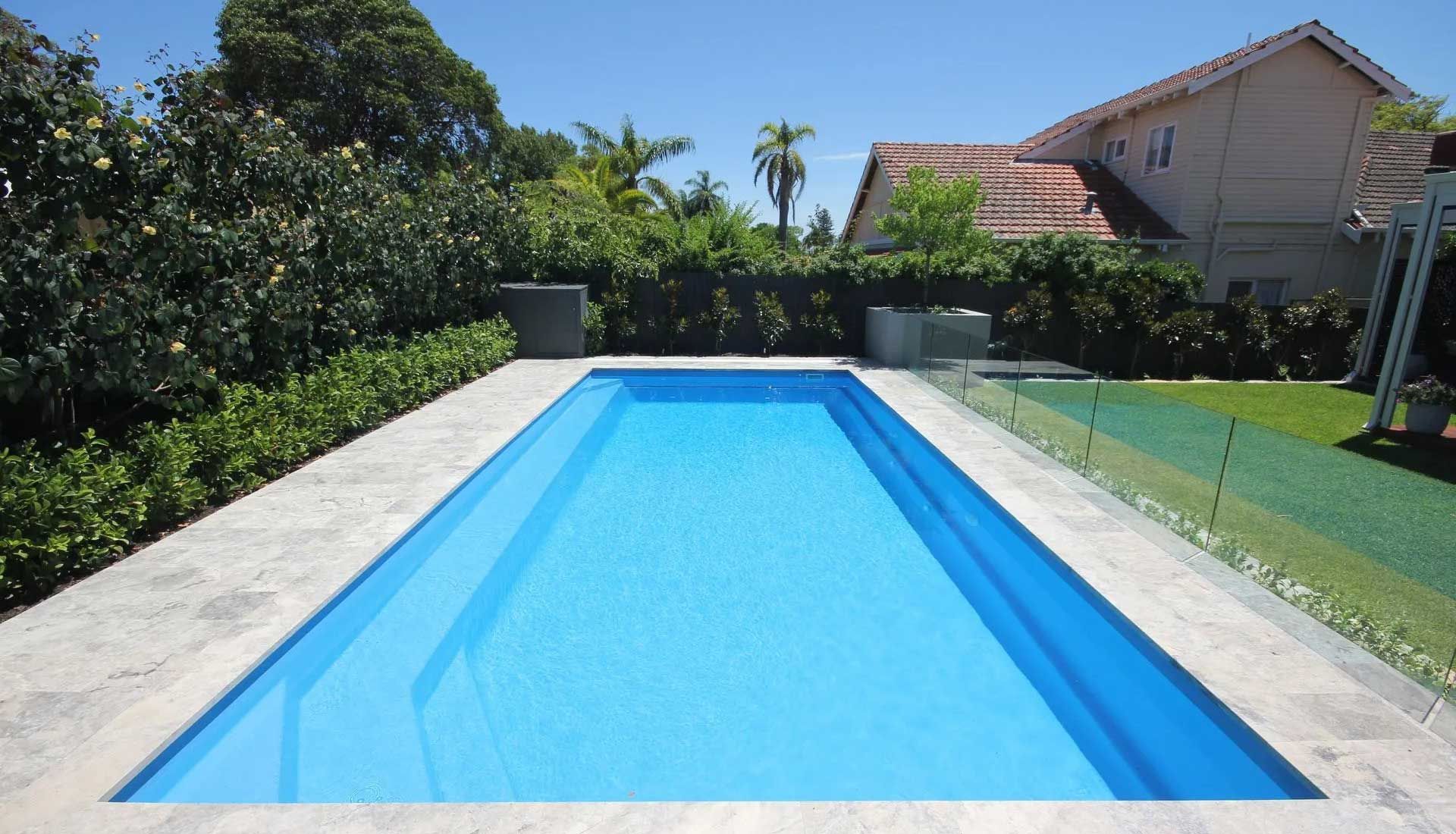 Rectangular blue swimming pool surrounded by stone tiles, with greenery and a house in the background.