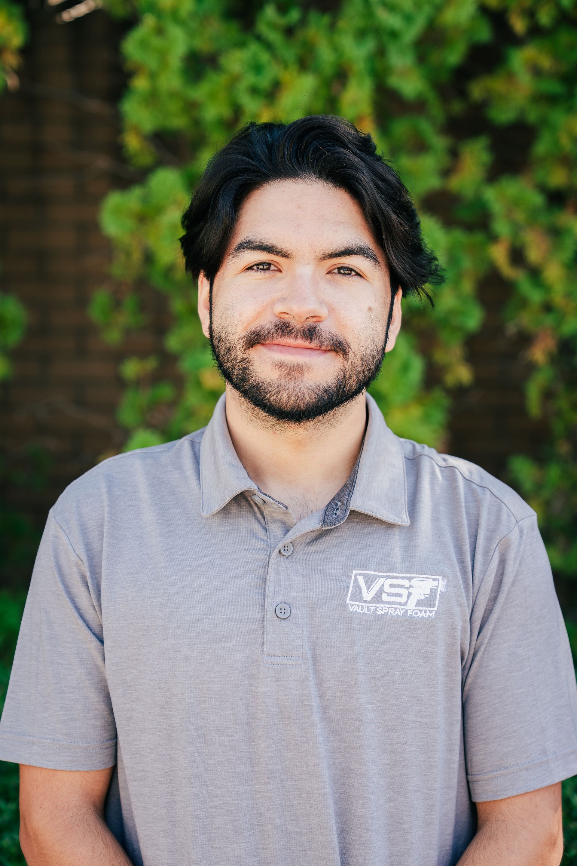 Portrait of a smiling person in a gray polo shirt standing outdoors against a green, blurred background