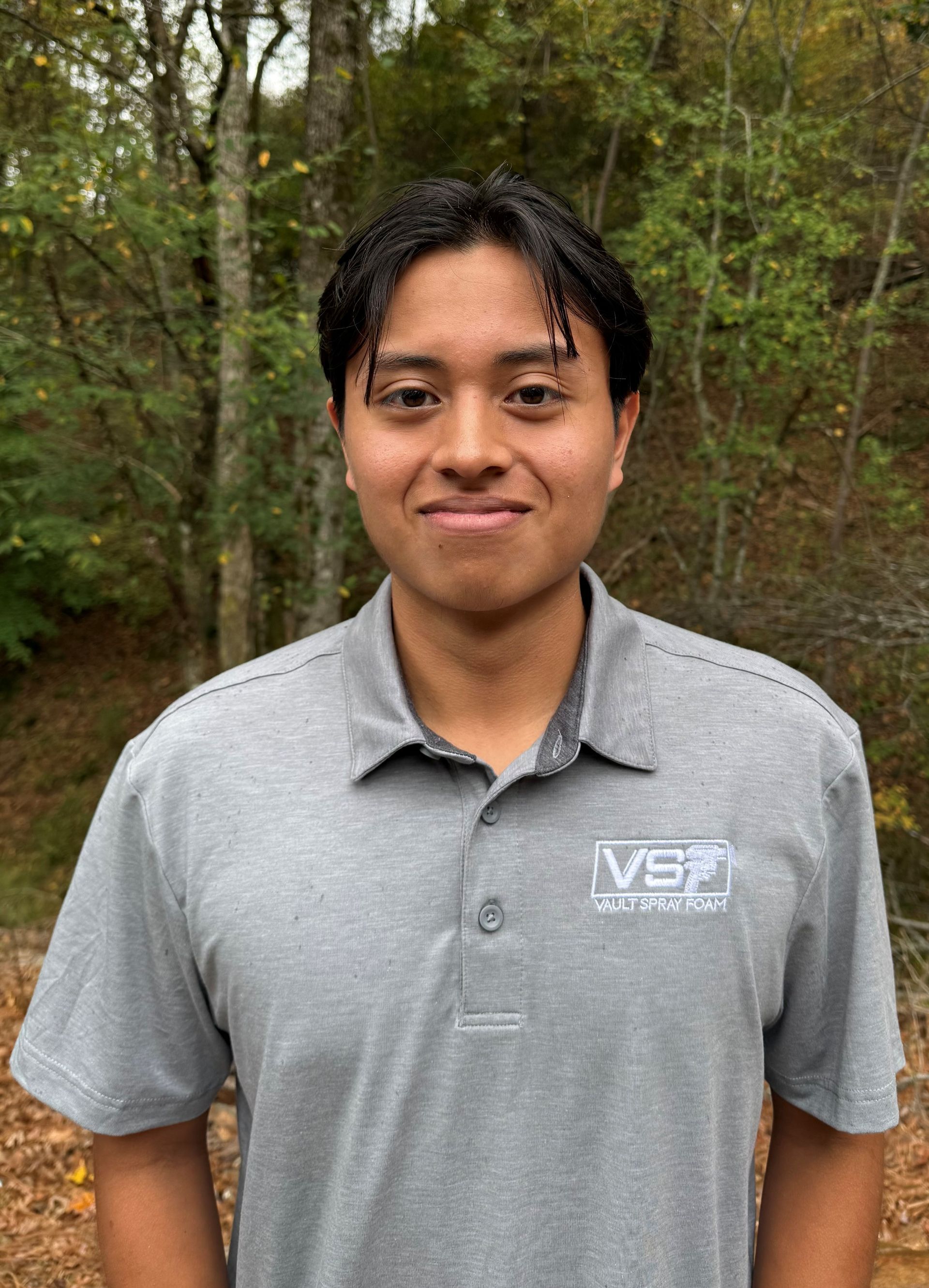 Person standing outdoors in a gray polo shirt, with trees and foliage in the background.