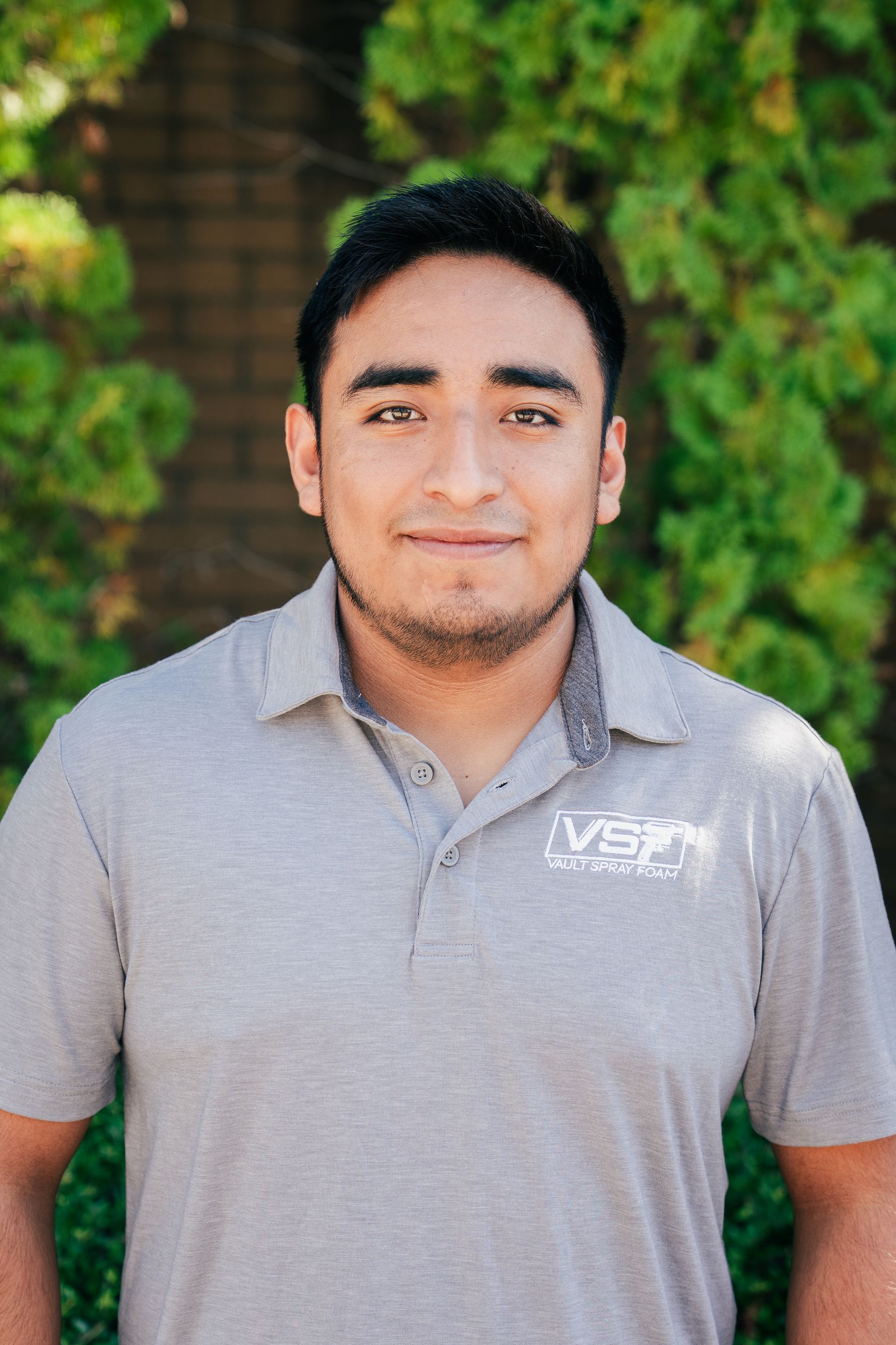 Man standing outdoors in a gray polo shirt, smiling slightly with green foliage in the background