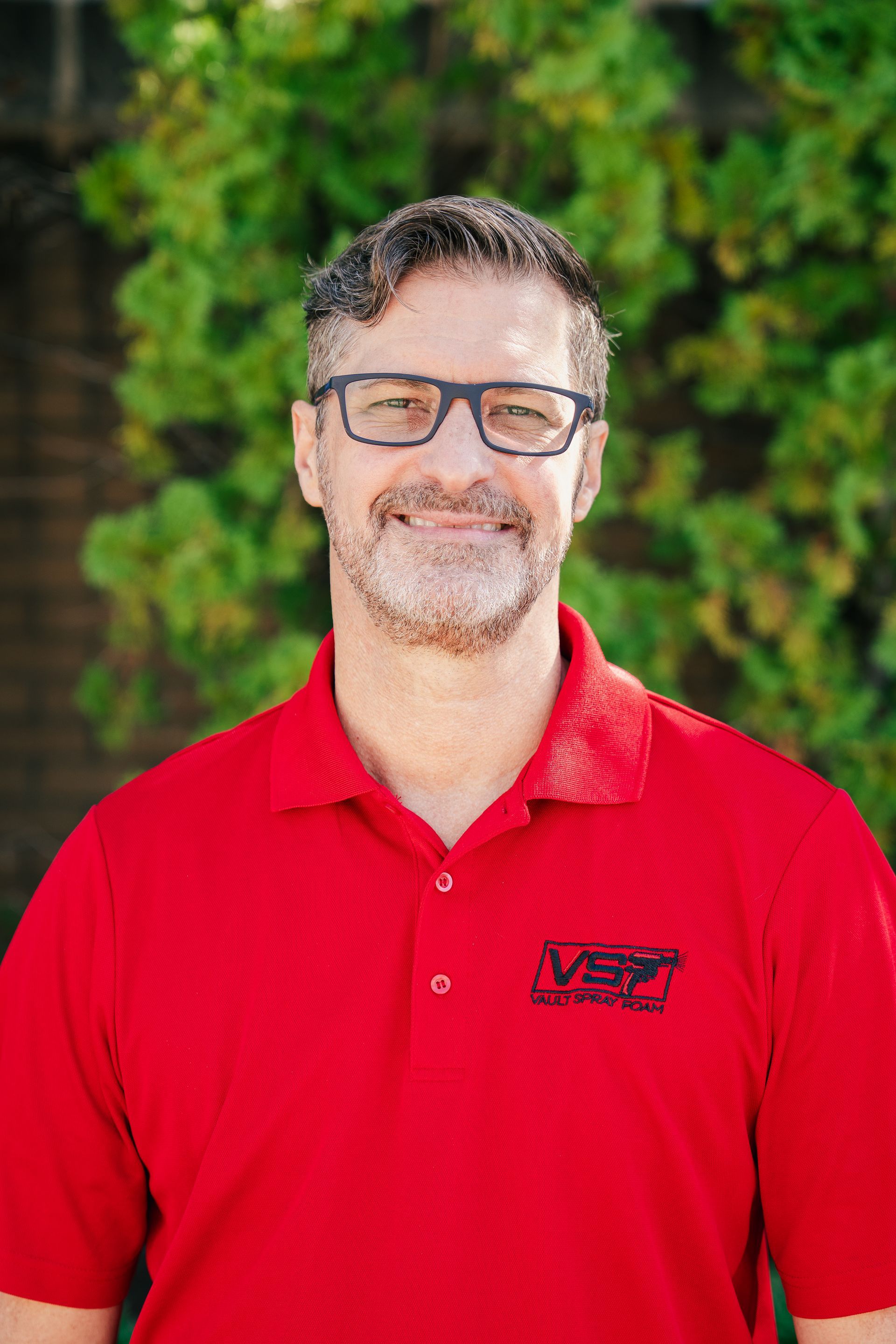 Smiling man in a red polo shirt and glasses standing outdoors in front of green foliage
