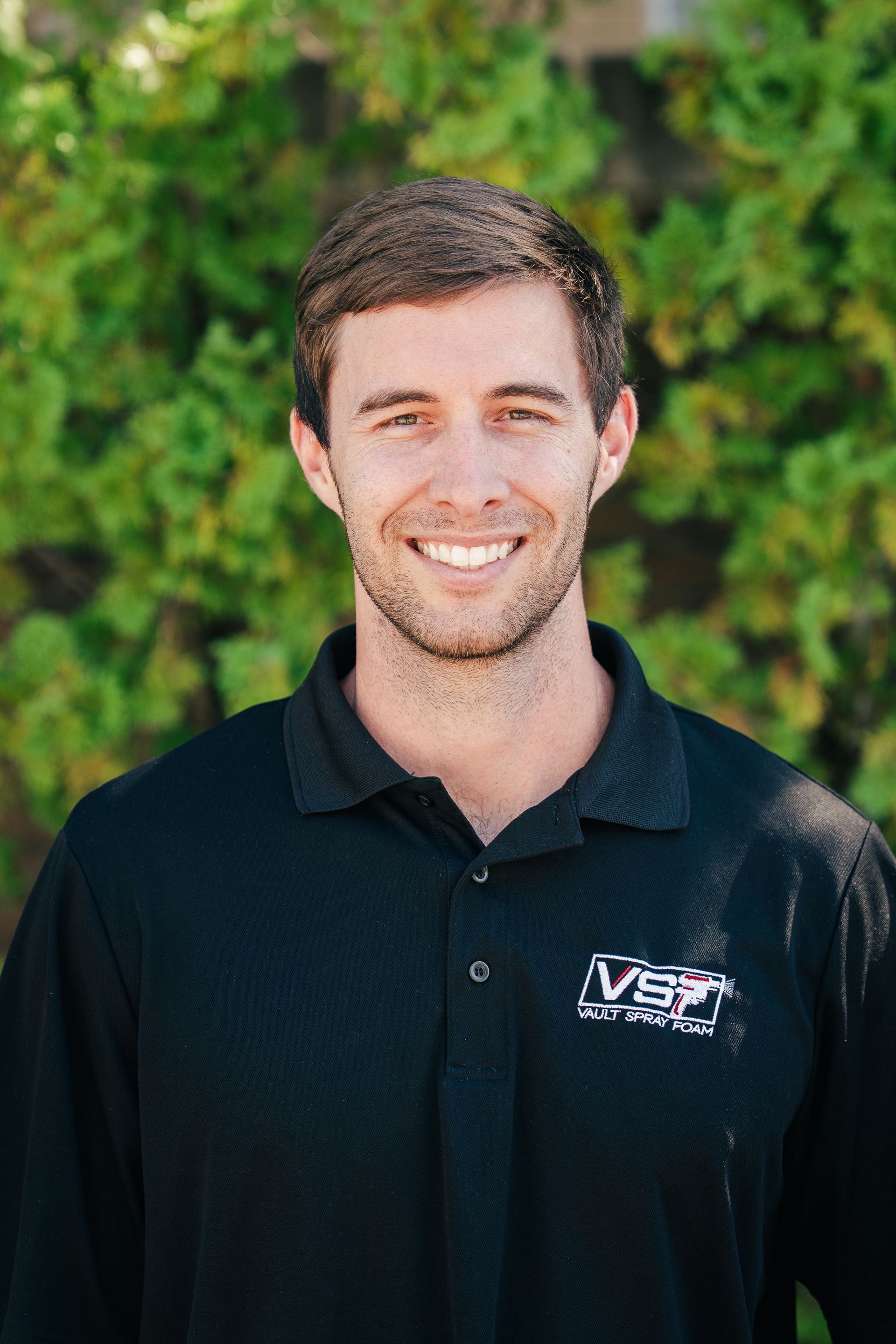 Smiling man in a black polo shirt standing outdoors against a green leafy background