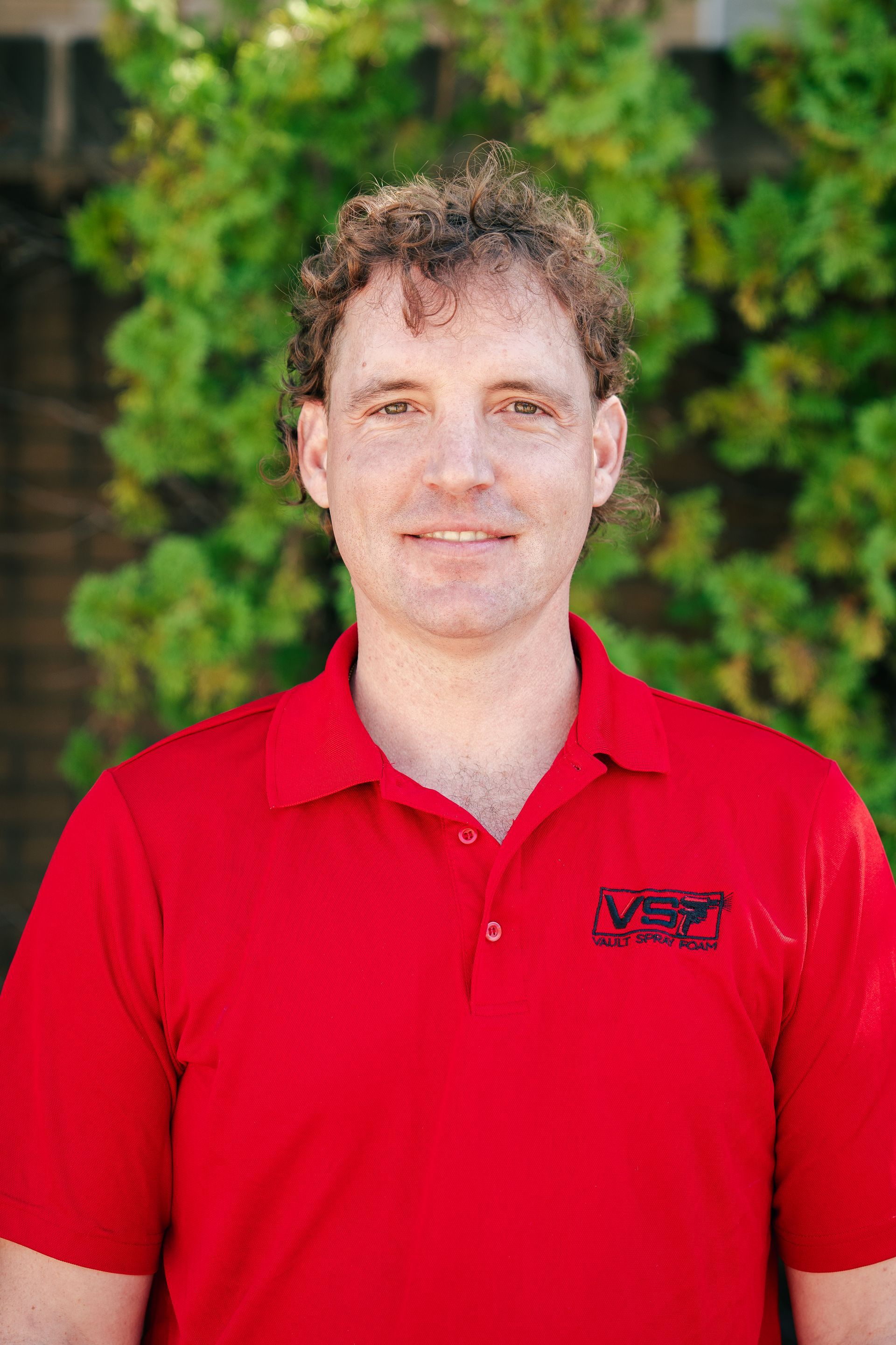 Man in a red polo shirt standing outdoors in front of green foliage