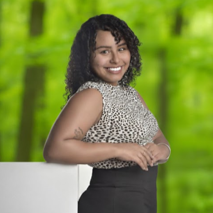 Smiling woman in a patterned top and black skirt, leaning on a white surface against a green blurred background