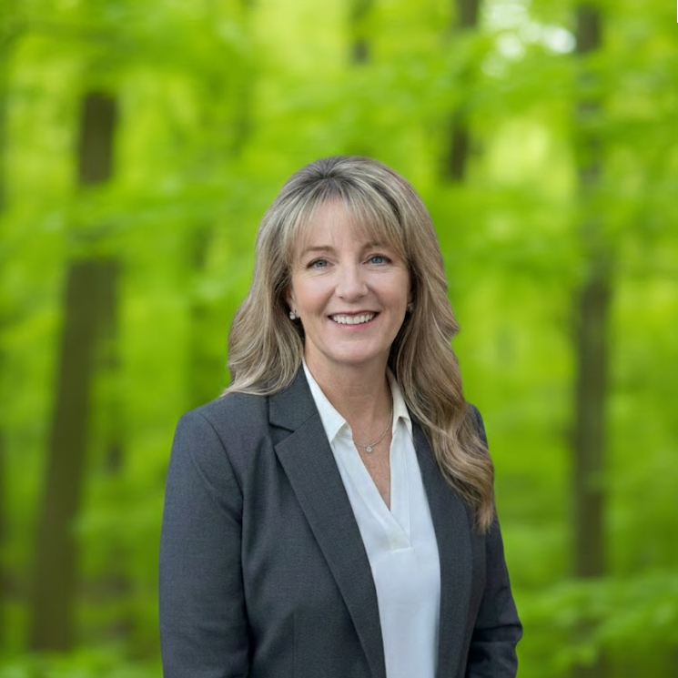 Smiling professional in a dark blazer and white blouse against a green forest background