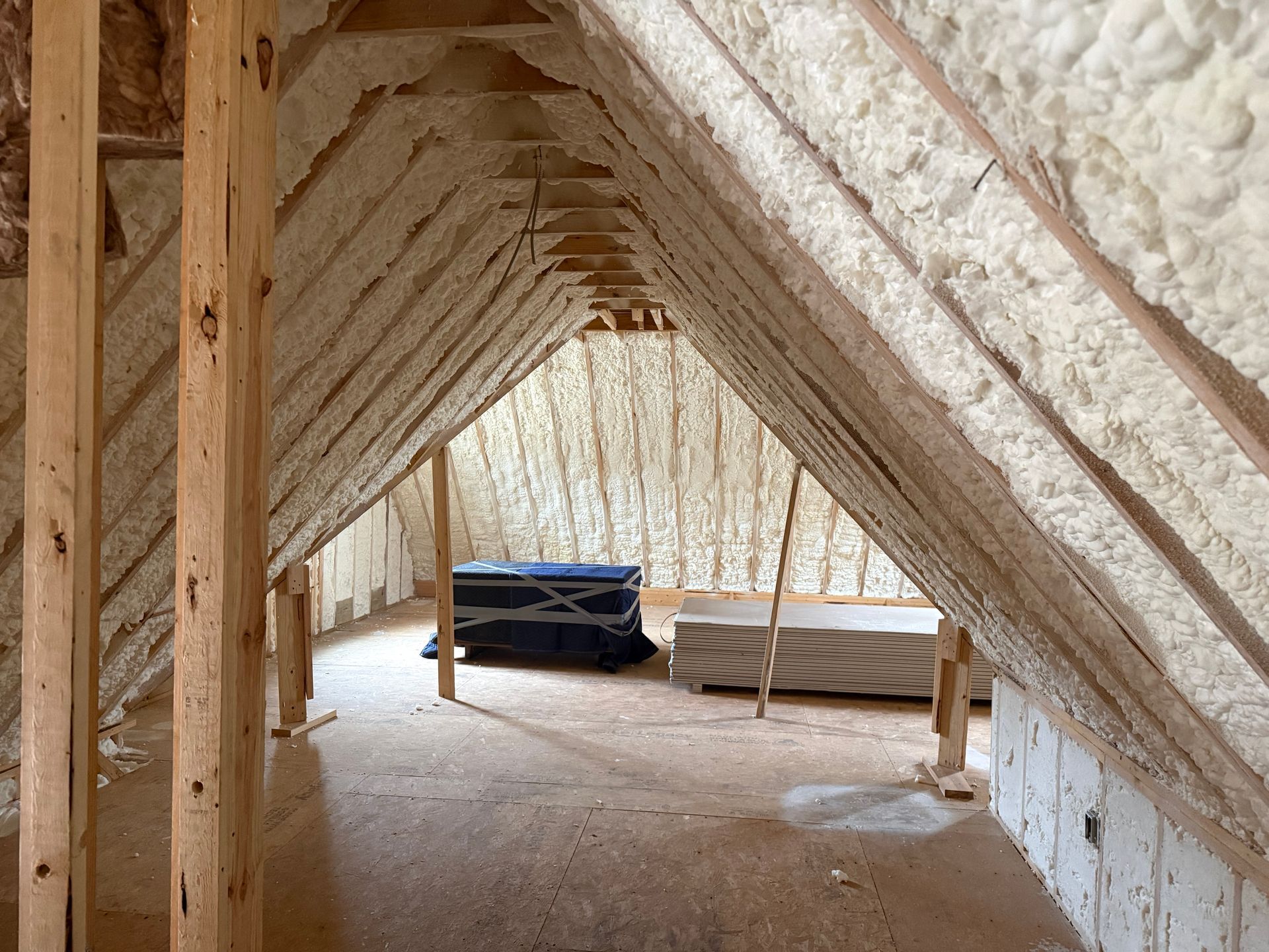 Unfinished attic with spray foam insulation, exposed rafters, and stacks of building materials on the floor