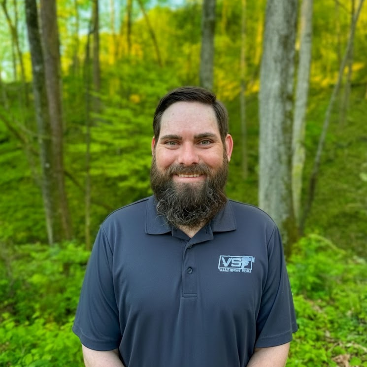 Smiling bearded man in a navy polo standing in a green wooded area