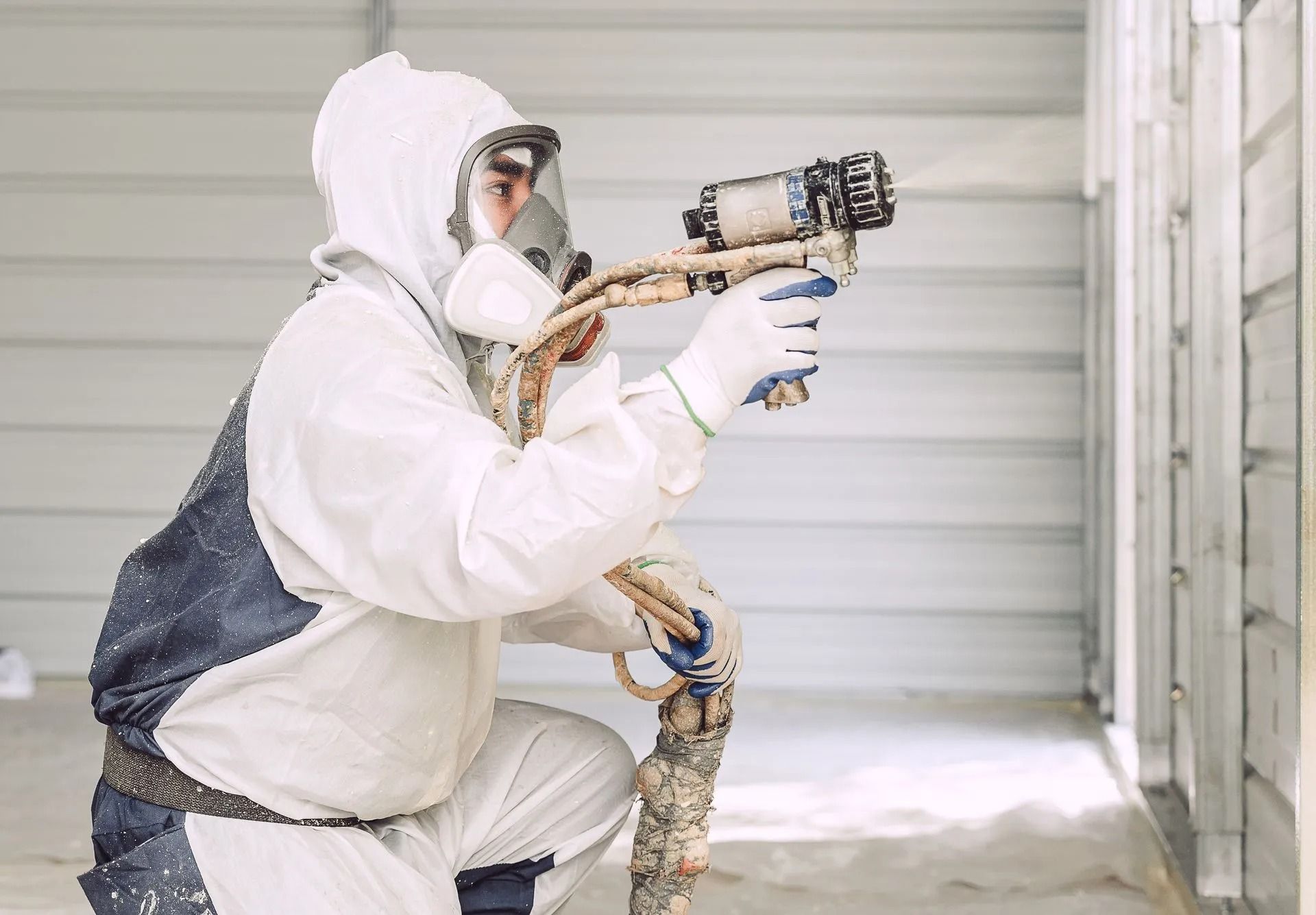 Worker in a white protective suit using a spray gun outdoors beside a metal wall.