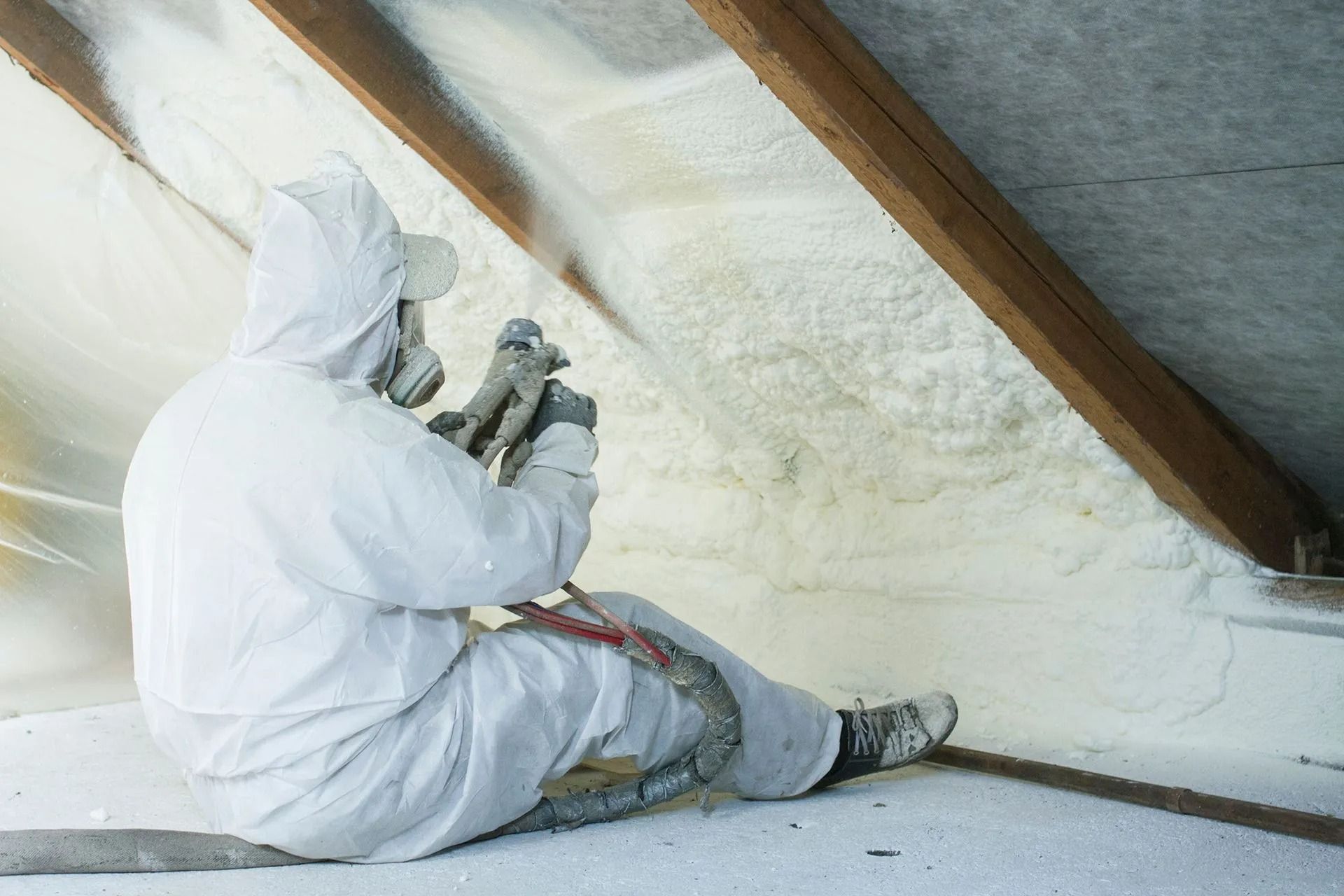 Worker in white protective suit spray-foaming insulation in an attic under a sloped roof