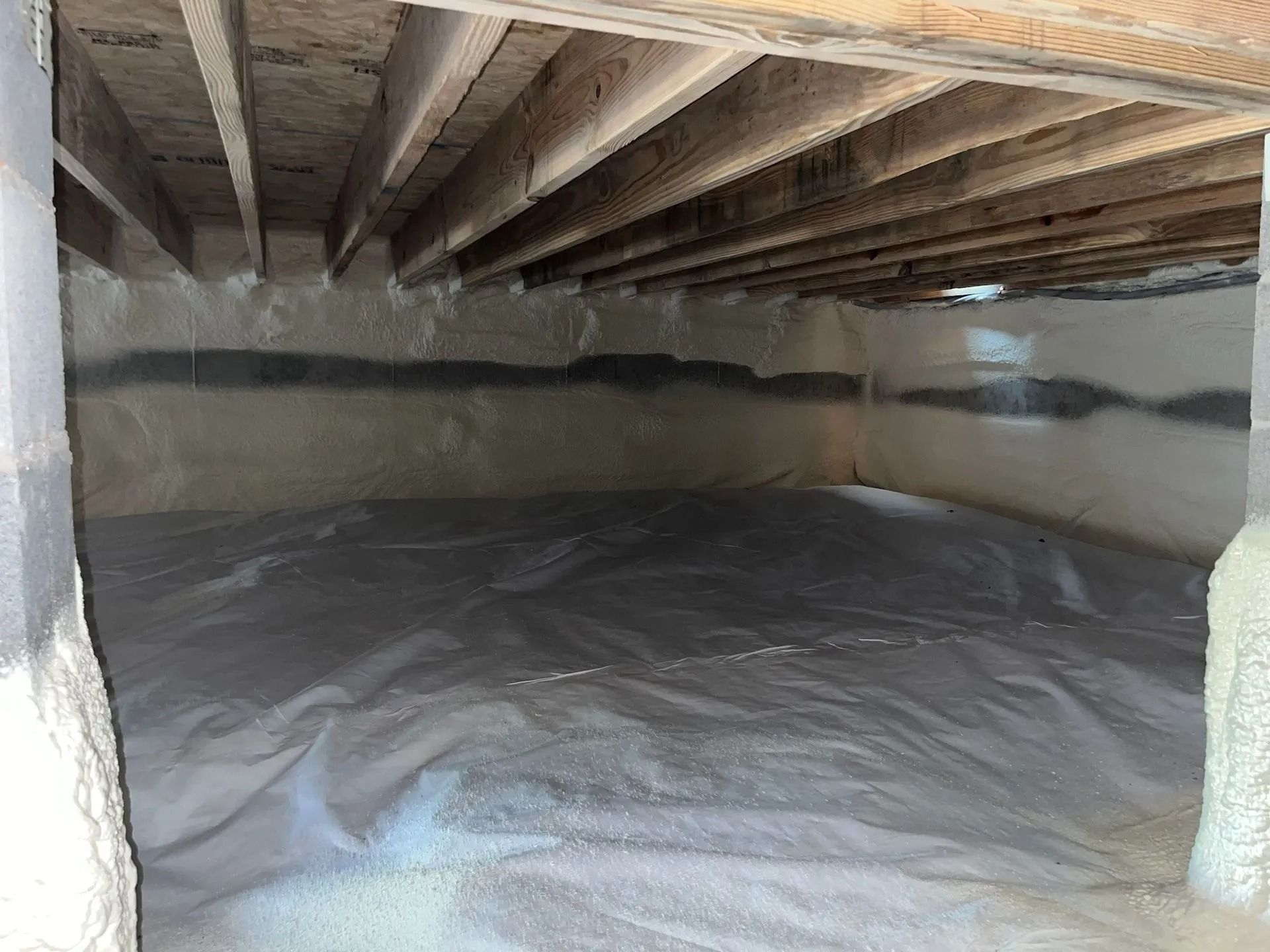 Crawlspace with exposed wooden joists, insulated walls, and a gray vapor barrier covering the floor.