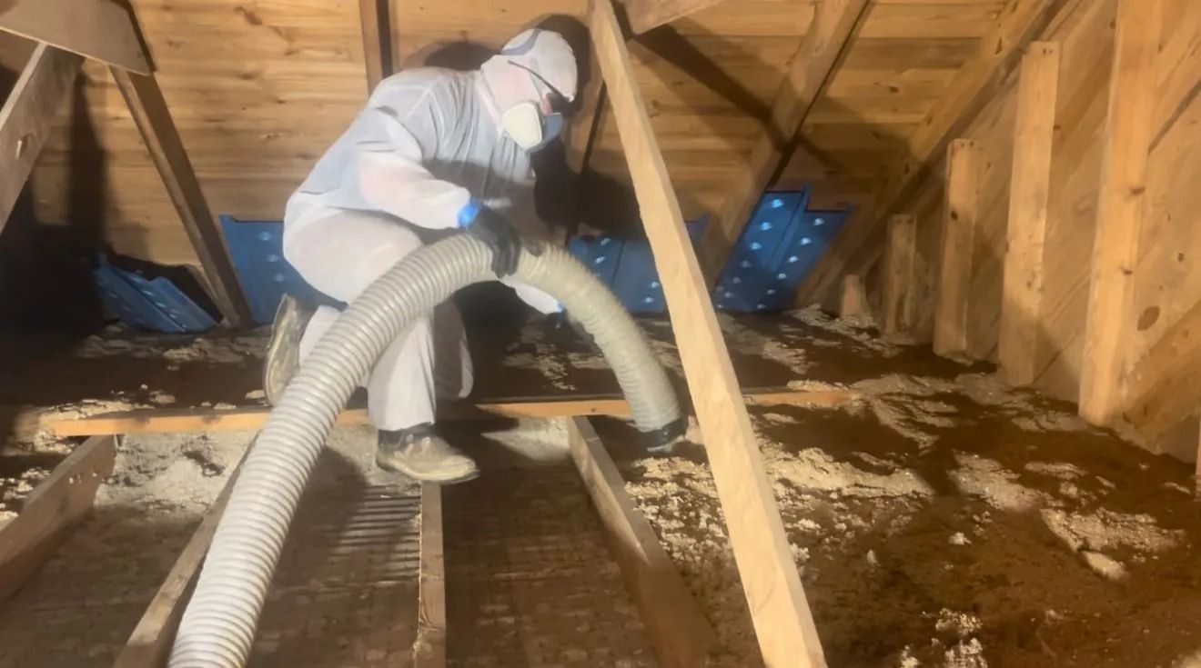 Worker in protective gear vacuuming insulation in a wooden attic crawlspace.