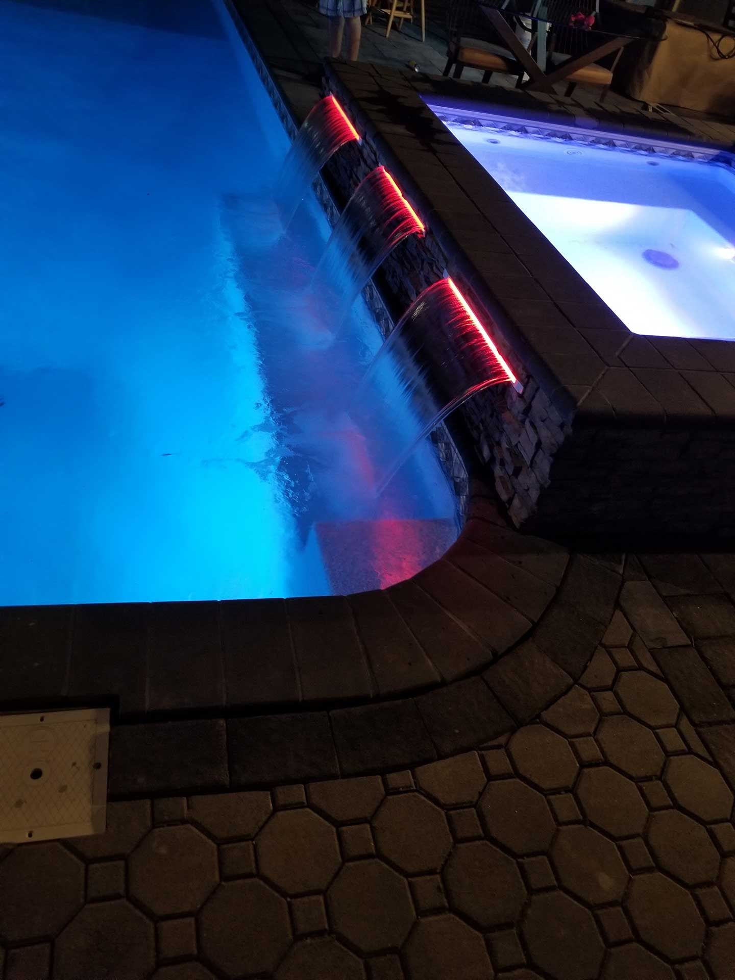 A swimming pool with a waterfall and lights on at night