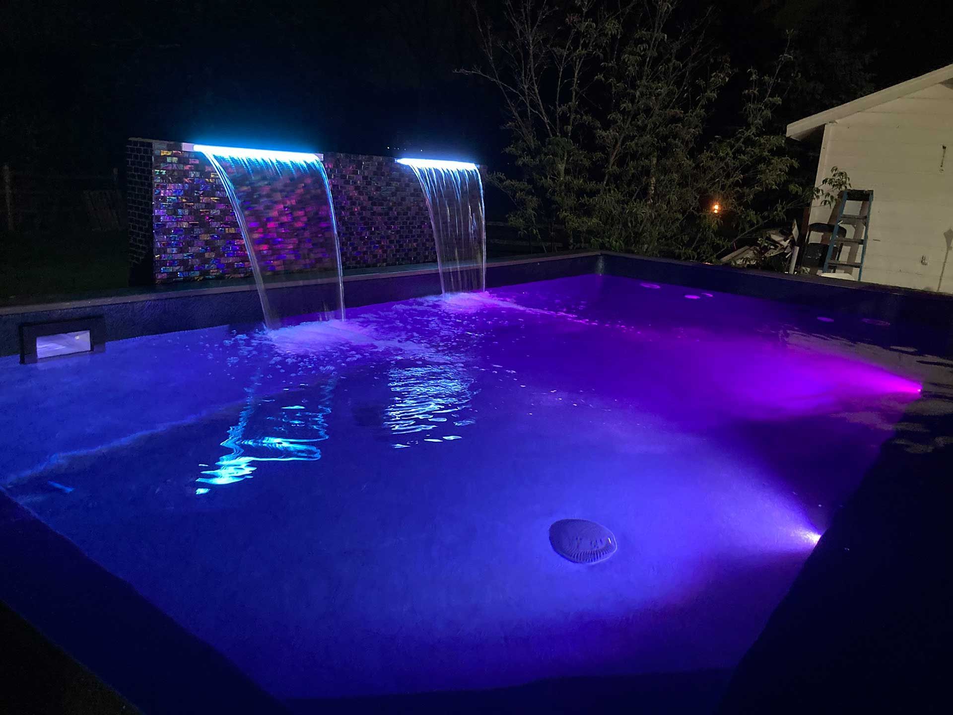 A swimming pool with purple lights and a waterfall at night