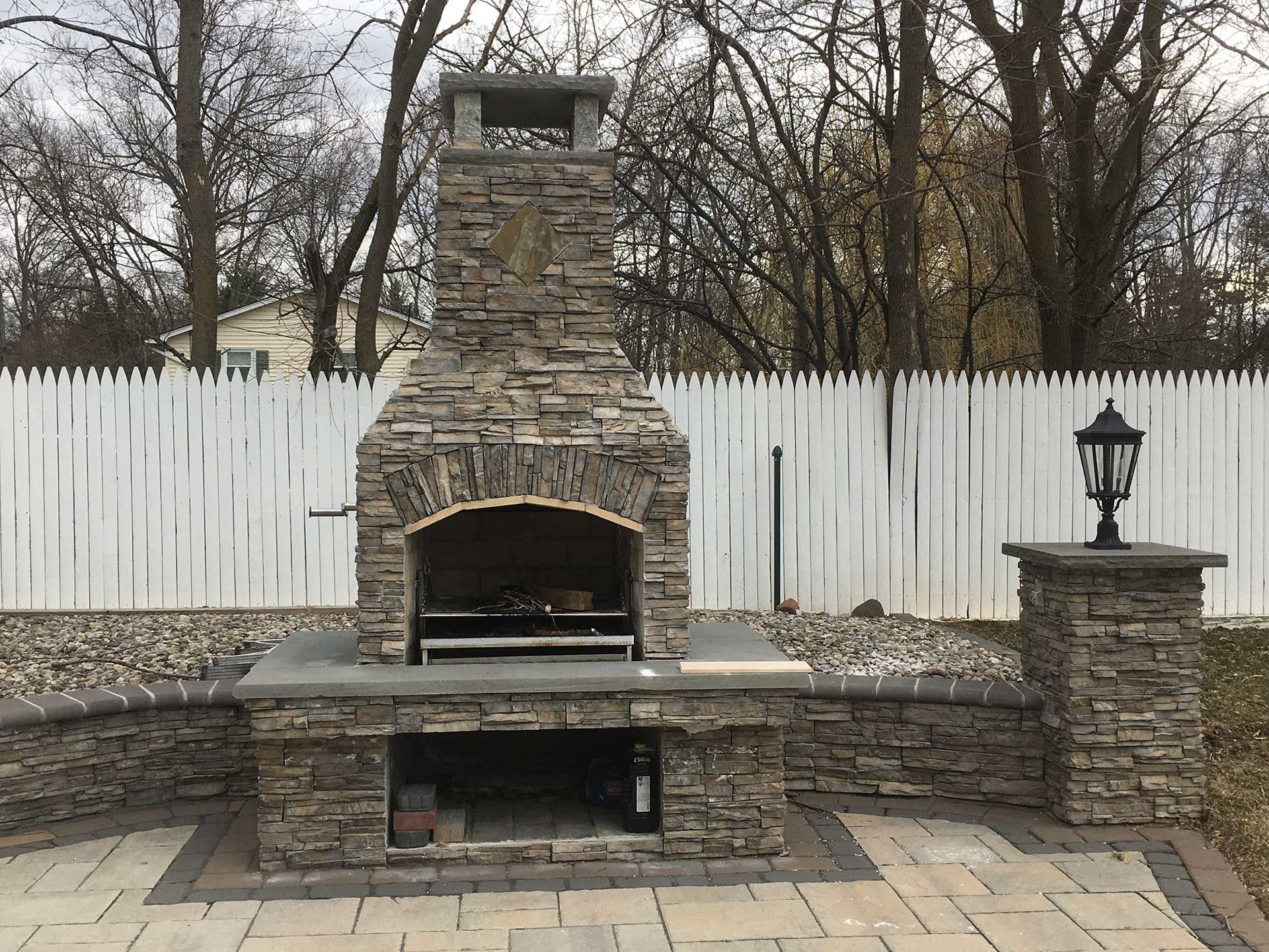 A stone fireplace is sitting on a patio next to a white picket fence