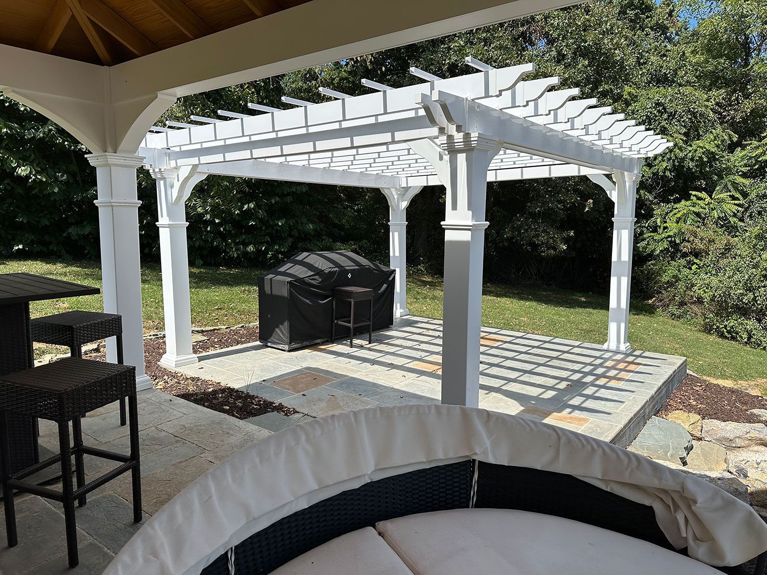 White pergola over a patio with seating, including a circular couch and bar stools. Green lawn in background.