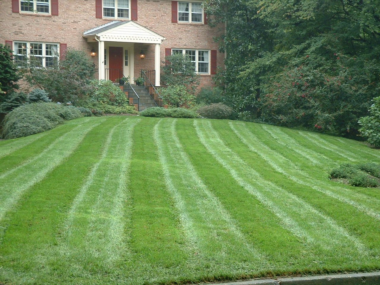 Lawn with striped patterns in front of a brick house with a covered porch and a red door.
