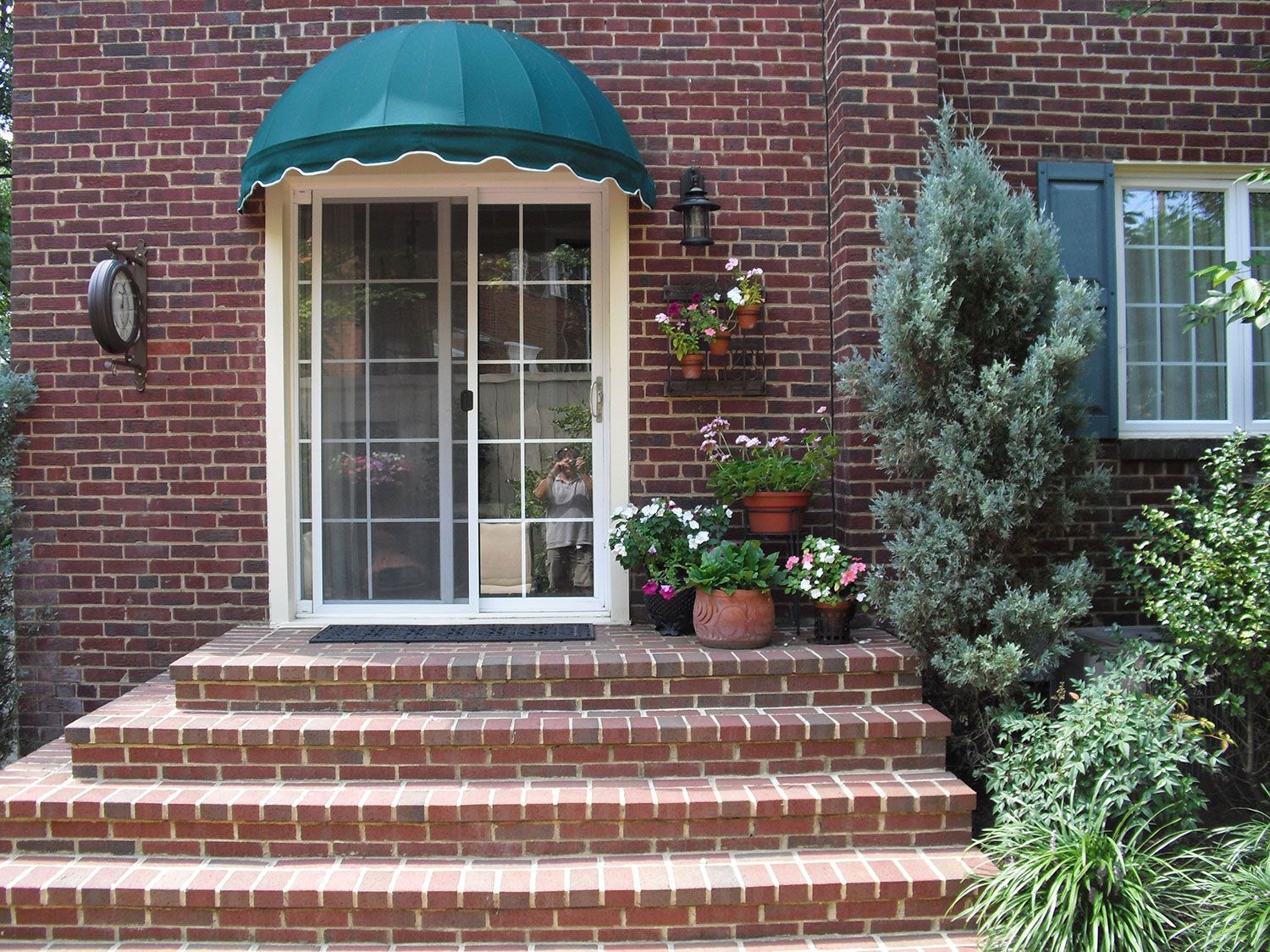 Brick house entrance with steps, screen door, awning, and potted flowers.