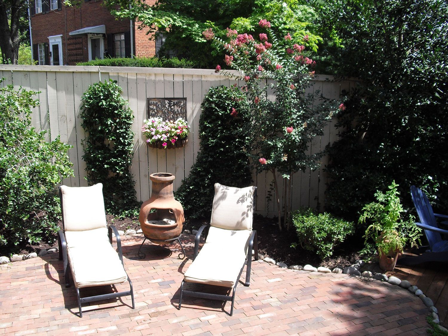 Two lounge chairs on a brick patio with a fire pit, surrounded by greenery and a fence.