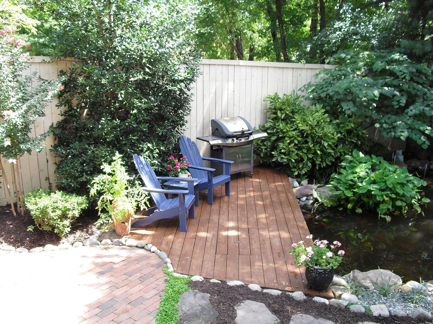 Two blue chairs and grill on a wooden deck next to a pond, surrounded by greenery and a fence.