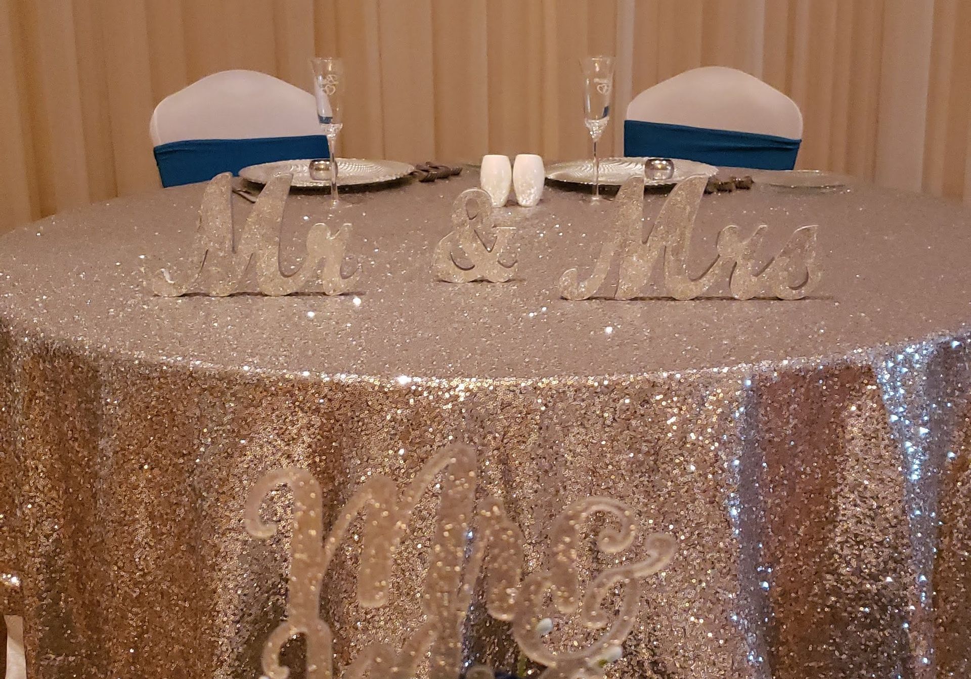 Sparkly silver table with “Mr & Mrs” decor, white chairs, and place settings at a wedding reception